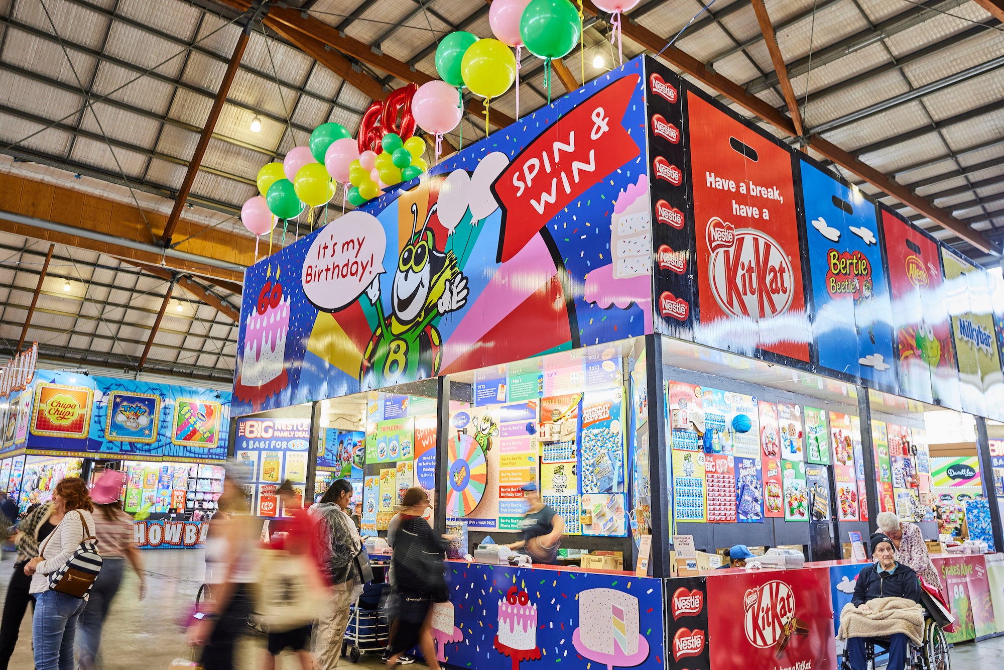 A colorful Bertie Beetle 60th birthday showbag booth inside a large indoor hall, with people walking around and balloons hanging above. Large signs read 'SPIN & WIN', 'It's my birthday!', and 'Have a break, save a KitKat', with KitKat branding prominently visible on the booth fronts.